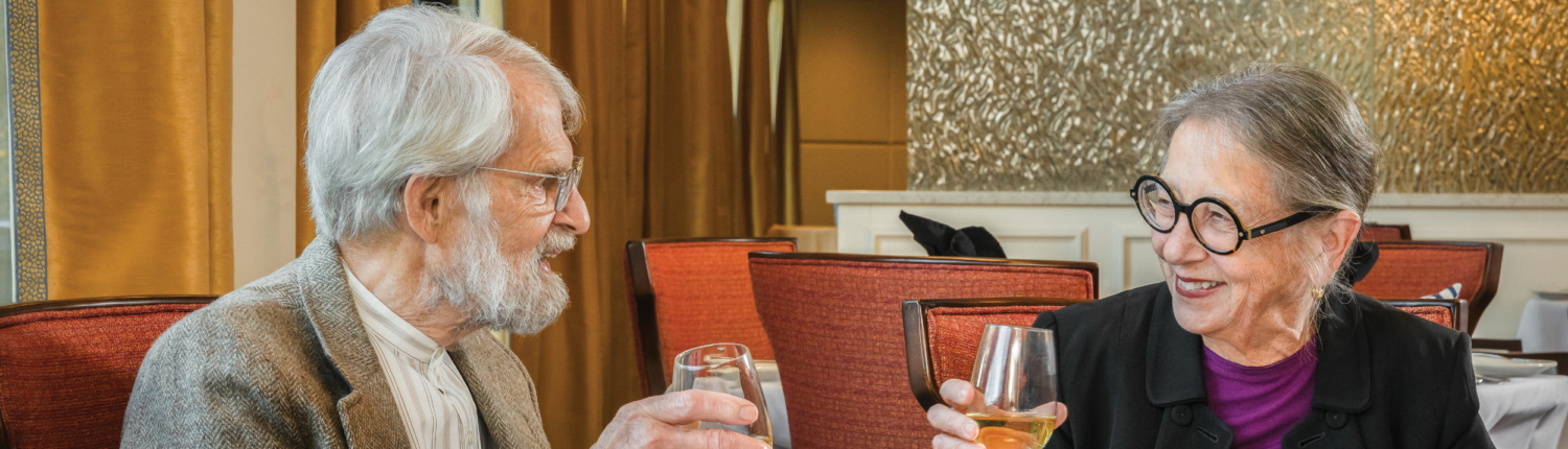 senior couple sitting at a dinner table holding glasses of white wine