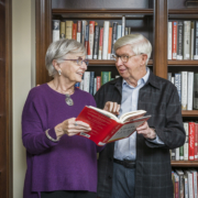 a senior couple standing in front of a bookcase reading a book together and talking