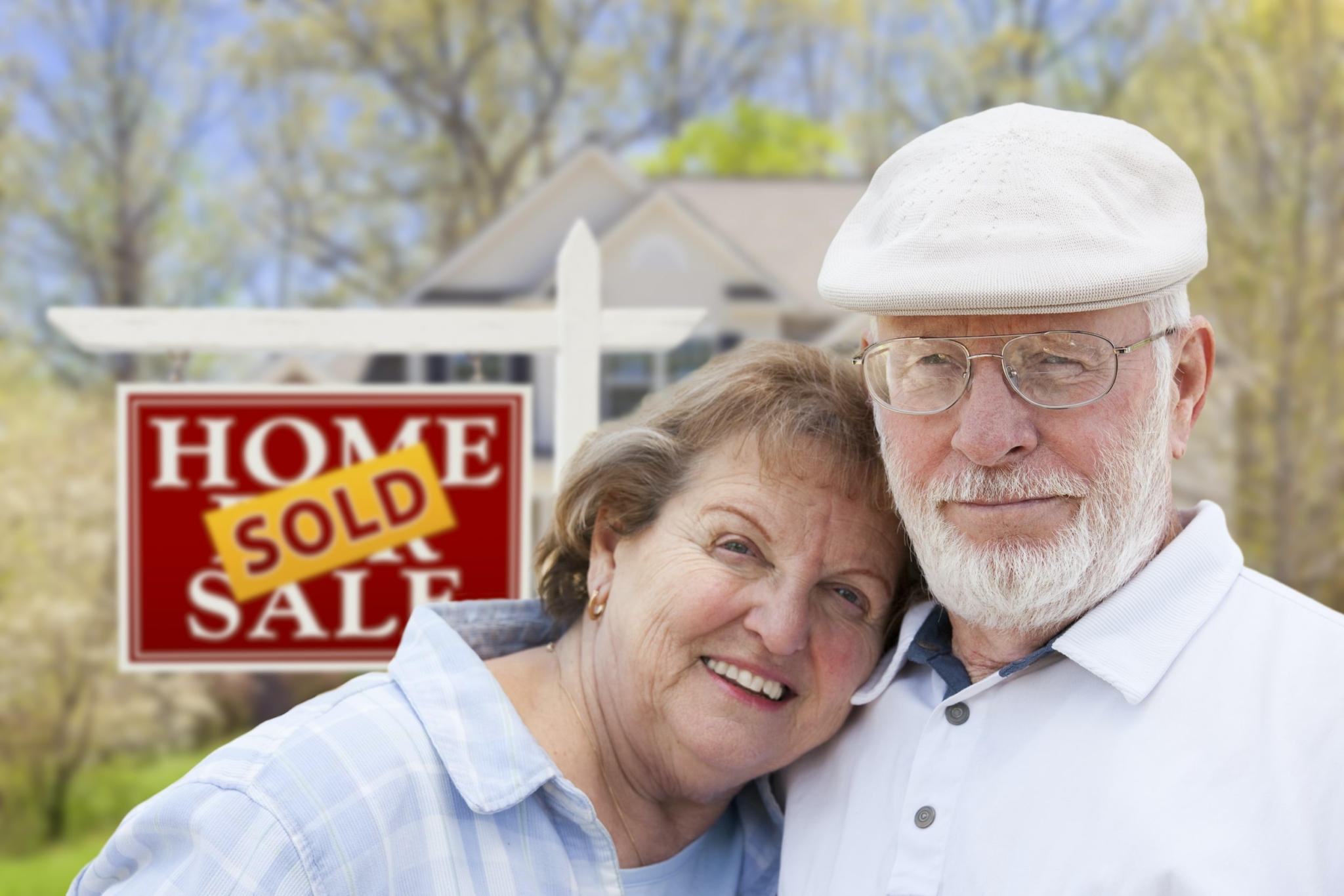 a senior couple standing in front of a "home for sale" sign with a SOLD sticker the sign. large home is in the background of the photo to illustrate that the couple is selling their home in order to downsize