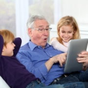 Senior man improving his tech skills on a tablet, sitting between two of his grandchildren on a couch.